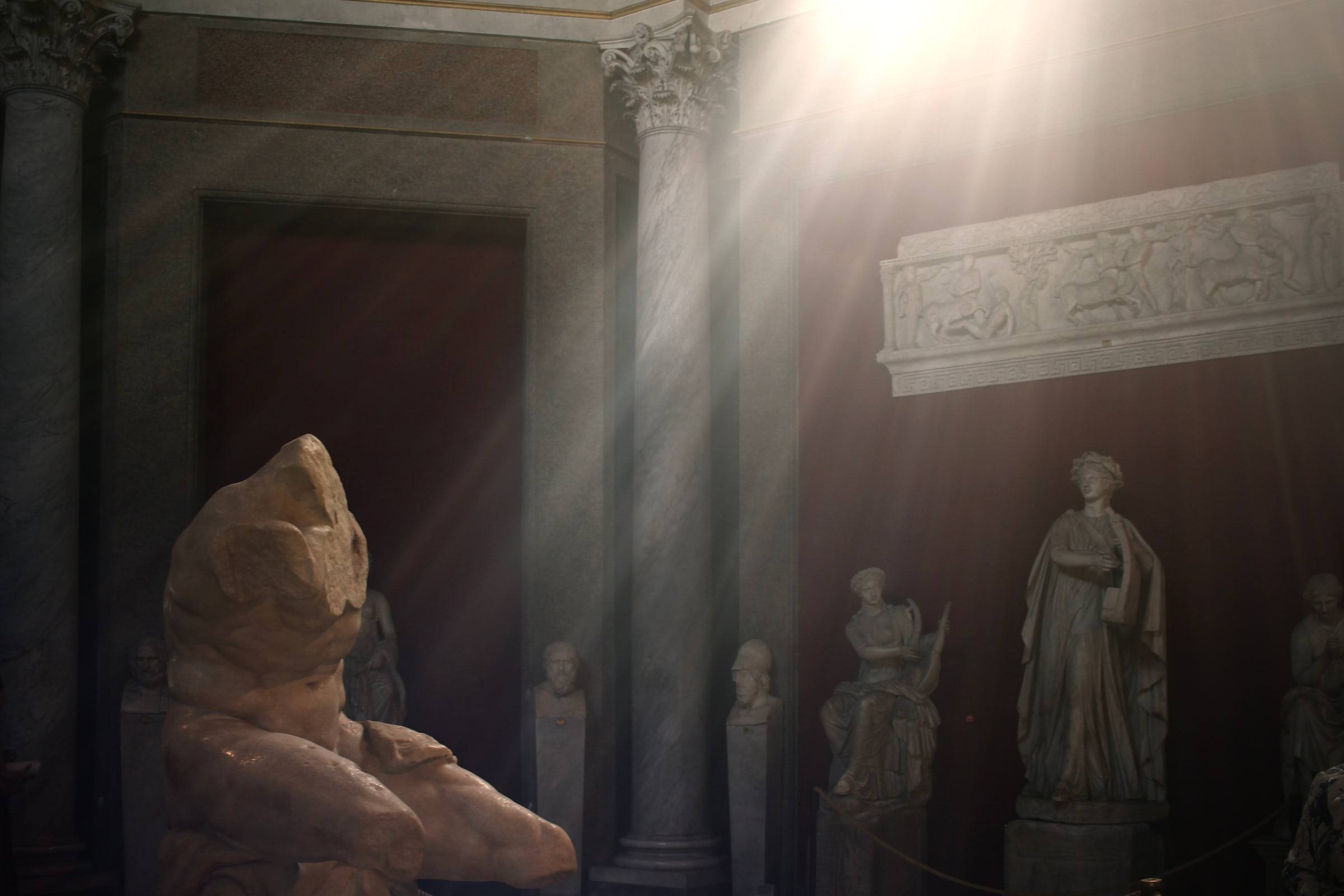 A shaft of daylight crosses a domed hall of Roman statues; a porphyry torso stands in shadow at the front.