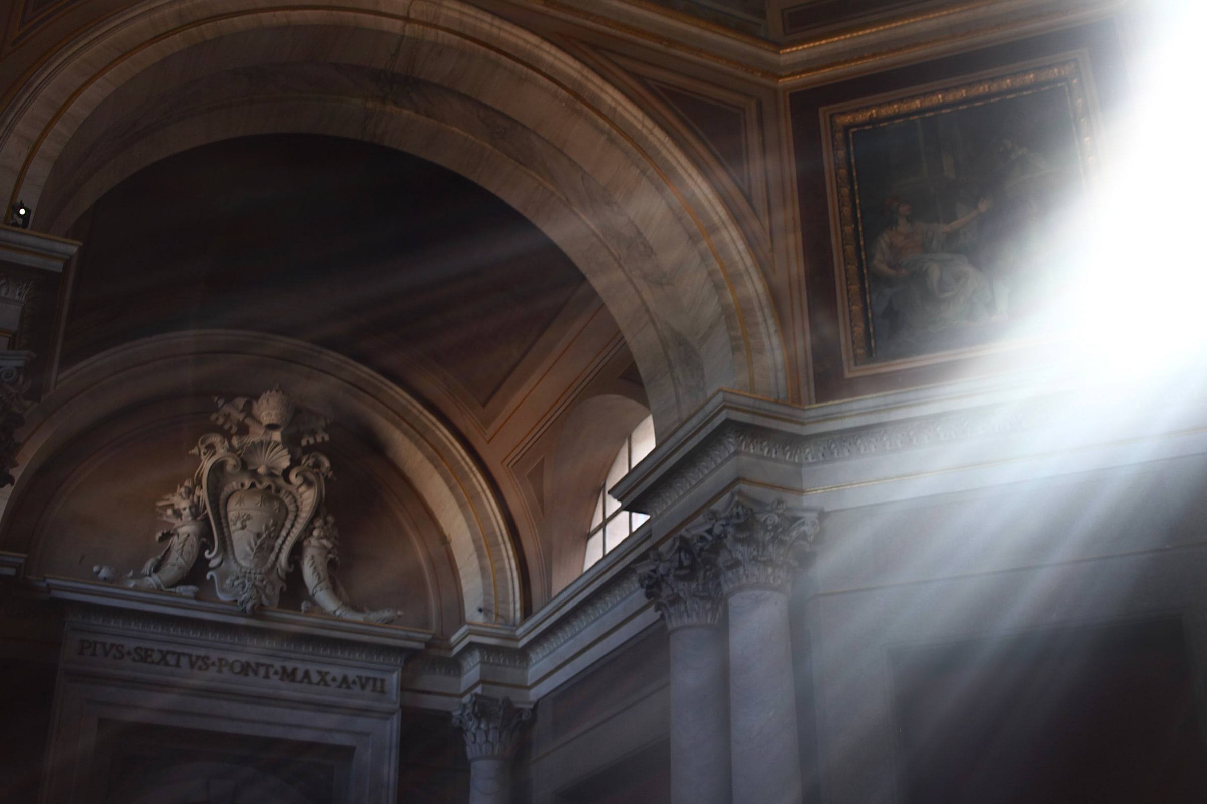 Sunlight entering a marble arch beneath an inscription to Pope Pius VI.